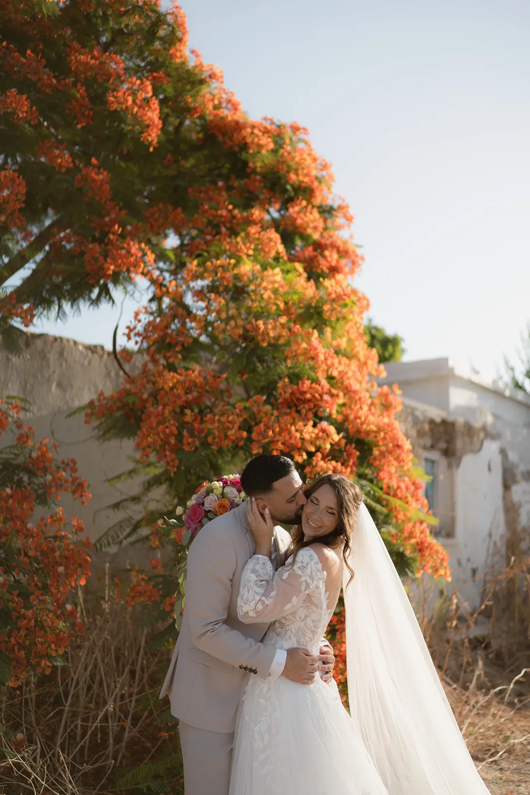 Bride and groom alone beneath a flame tree in full orange bloom beside an old stone building in Cyprus during their elopement portraits.