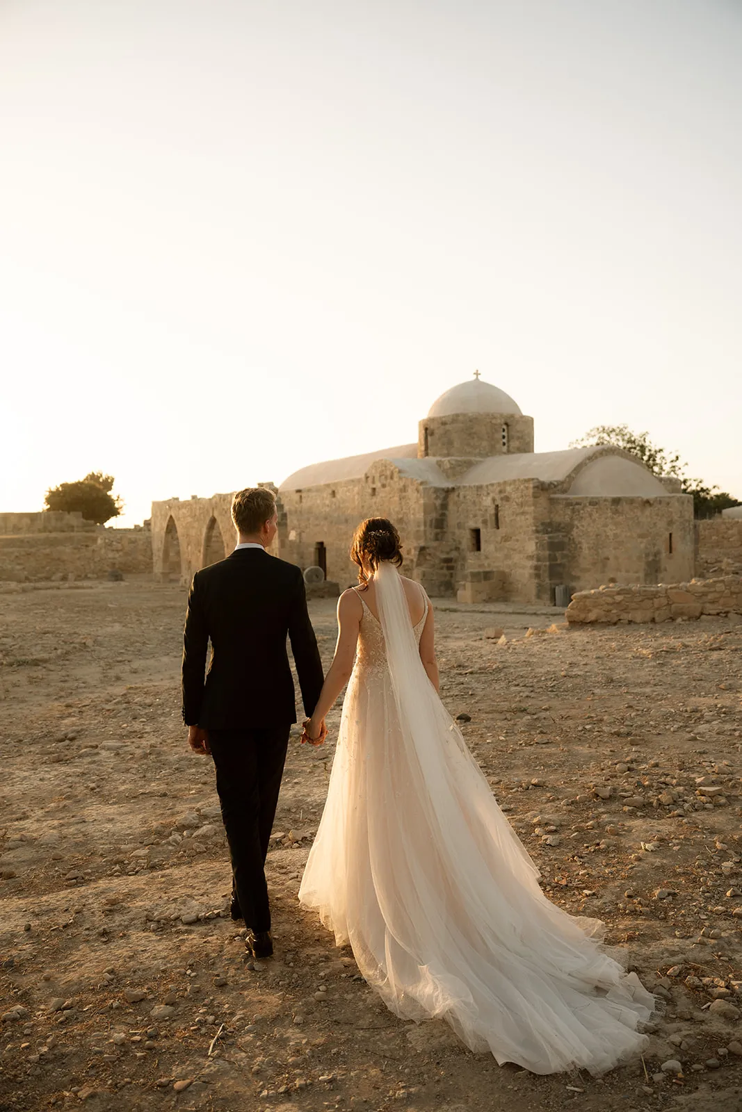 Couple walking hand in hand towards a Byzantine stone church at golden hour in the Paphos countryside, Cyprus, with nobody else in sight.