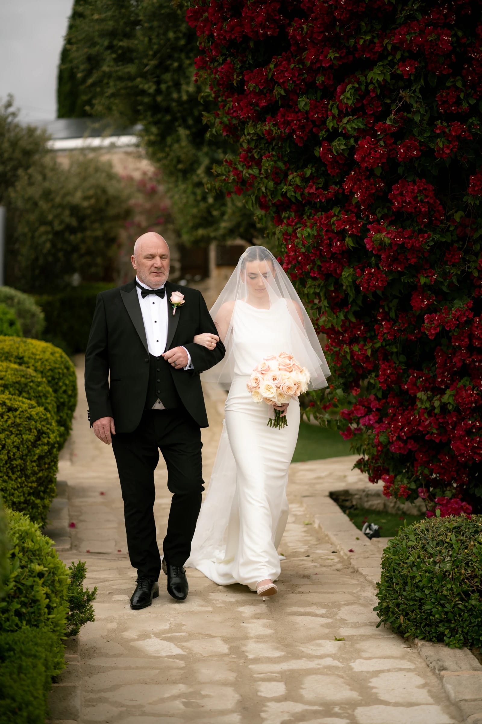 Bailey walking down the aisle with her father past red bougainvillea at Liopetro, Paphos.