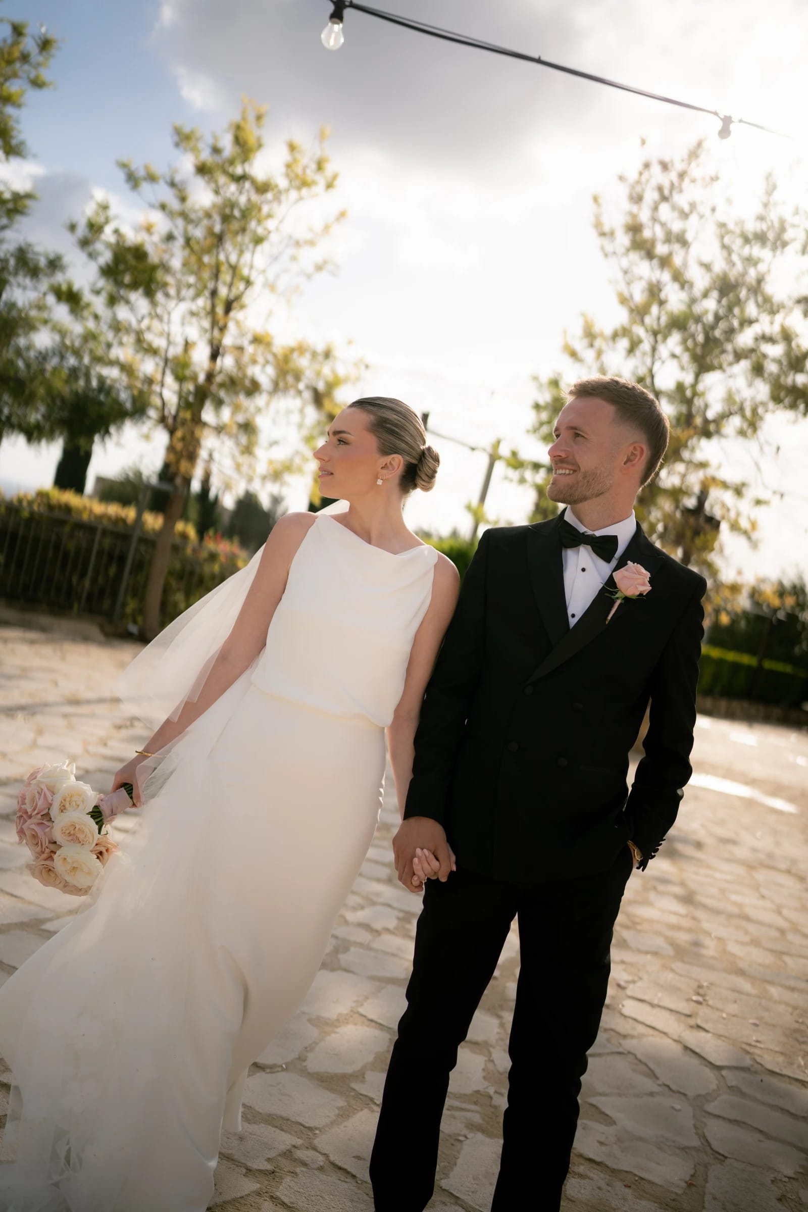 Daniel and Bailey walking hand in hand under the Liopetro string lights in golden hour, Kouklia, Paphos.