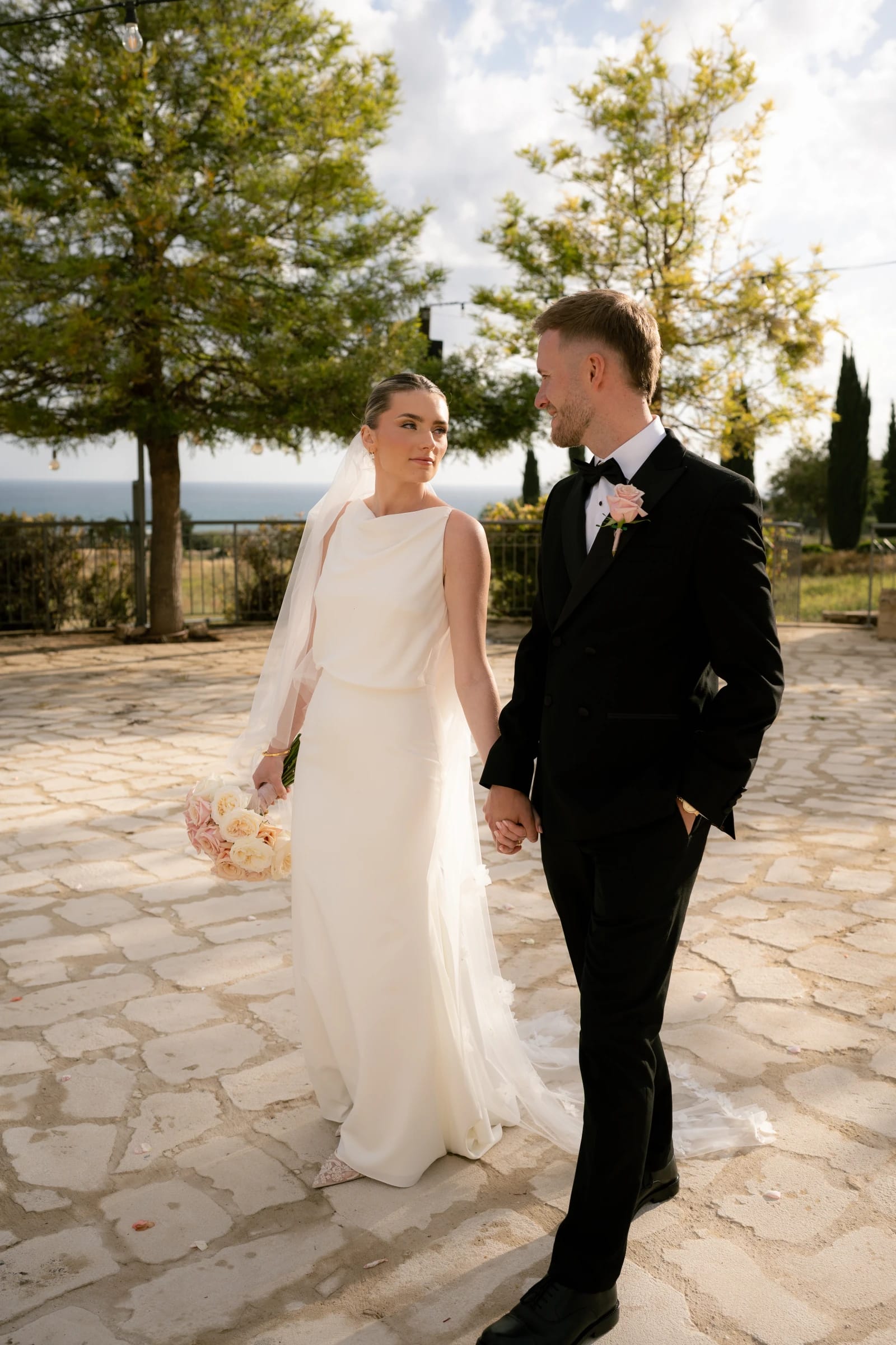 Daniel and Bailey hand in hand on the Liopetro courtyard stone with the Paphos coast behind them.