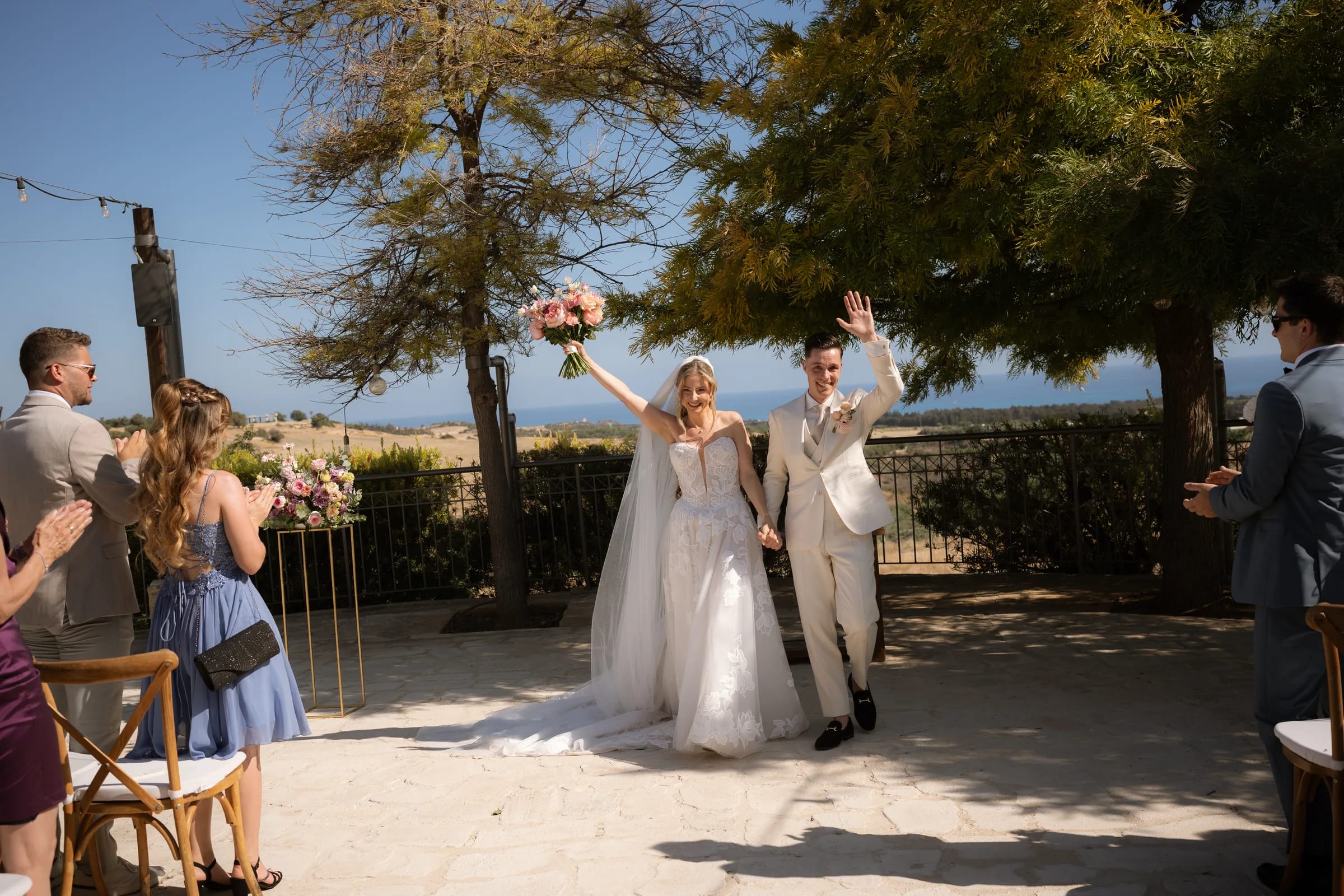 Frank and Franziska walking back up the aisle after their ceremony — bouquet raised and groom waving — as guests applaud, with the sea and Paphos countryside behind, at Liopetro, Kouklia.