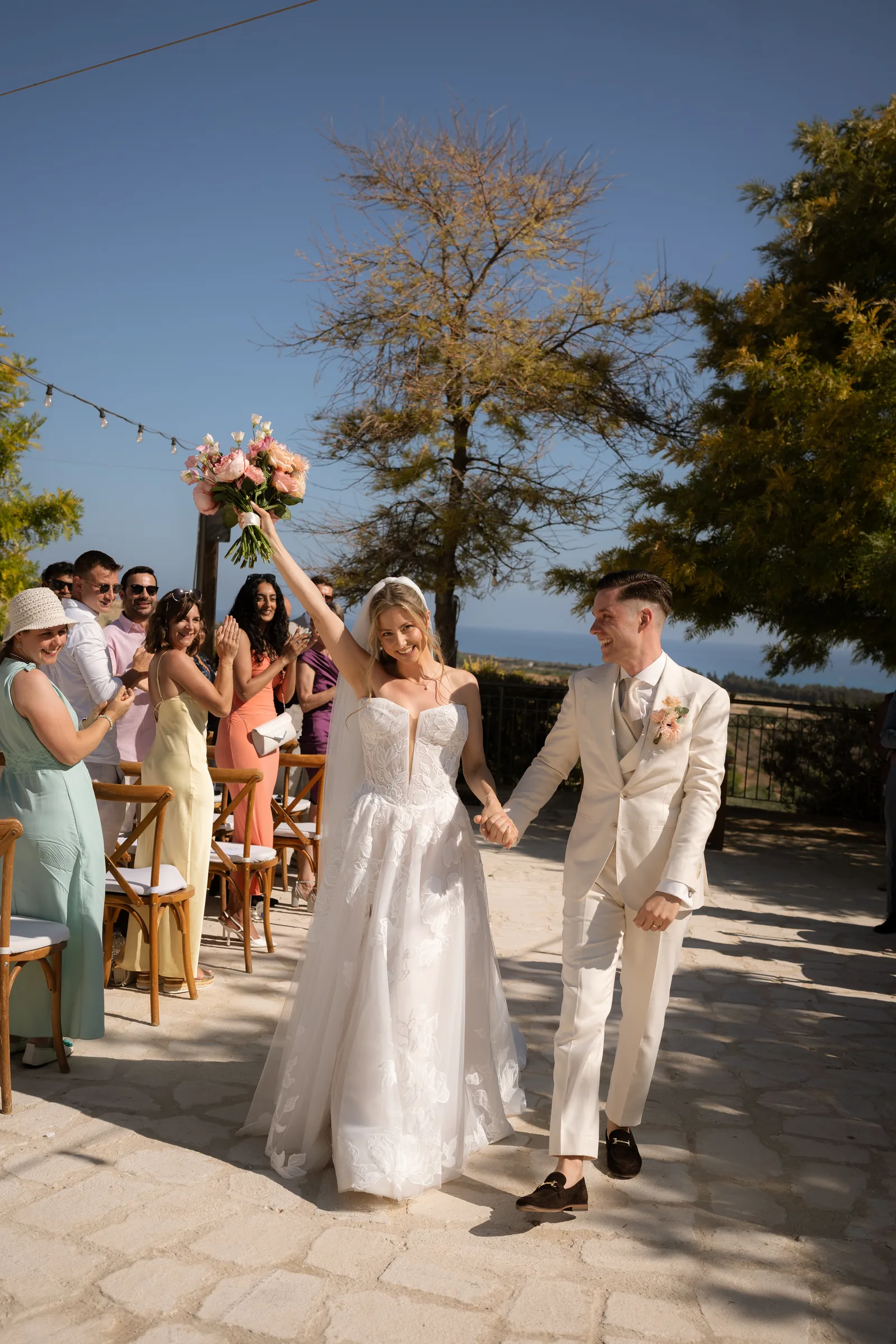 Franziska raising her bouquet high as she and Frank walk back down the aisle to applause after their ceremony at Liopetro, Kouklia, Paphos.