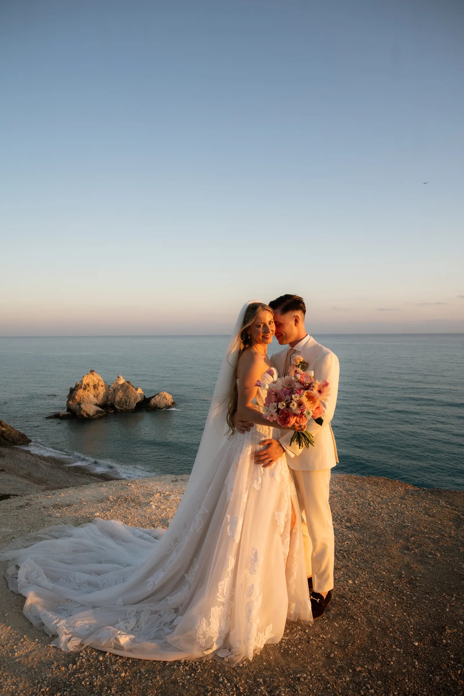 Frank and Franziska touching foreheads in warm golden light on the clifftop at Aphrodite's Rock, the sea rock behind them, near Paphos, Cyprus.