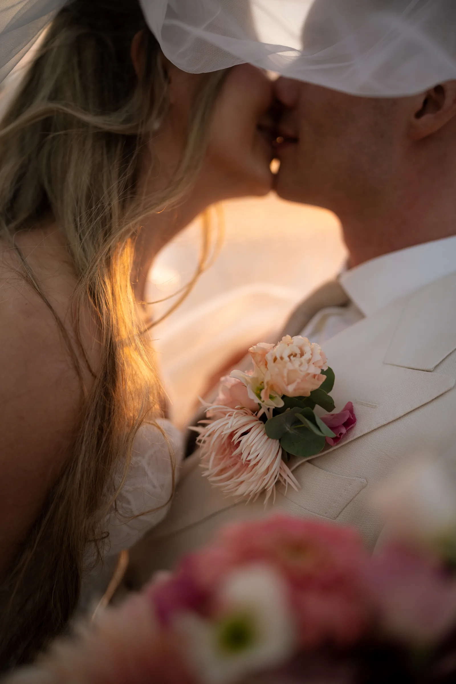 Frank and Franziska kissing beneath her veil at sunset, his coral boutonnière catching the light, at Aphrodite's Rock, Paphos.
