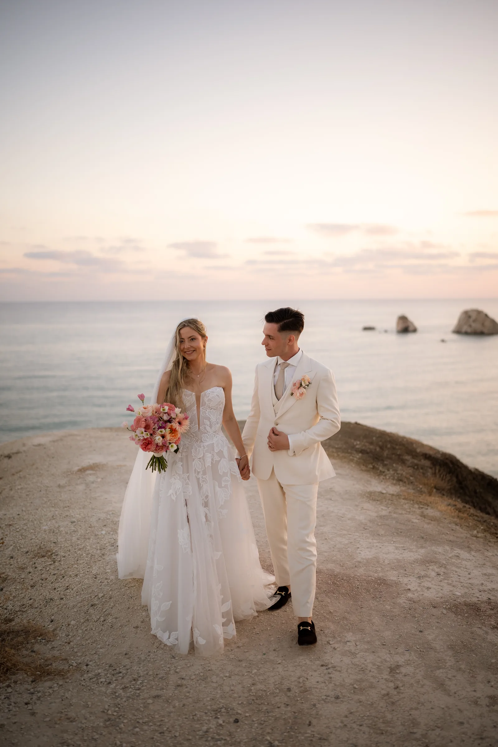 Frank and Franziska walking hand in hand with her coral bouquet at Aphrodite's Rock at sunset, Paphos, Cyprus.