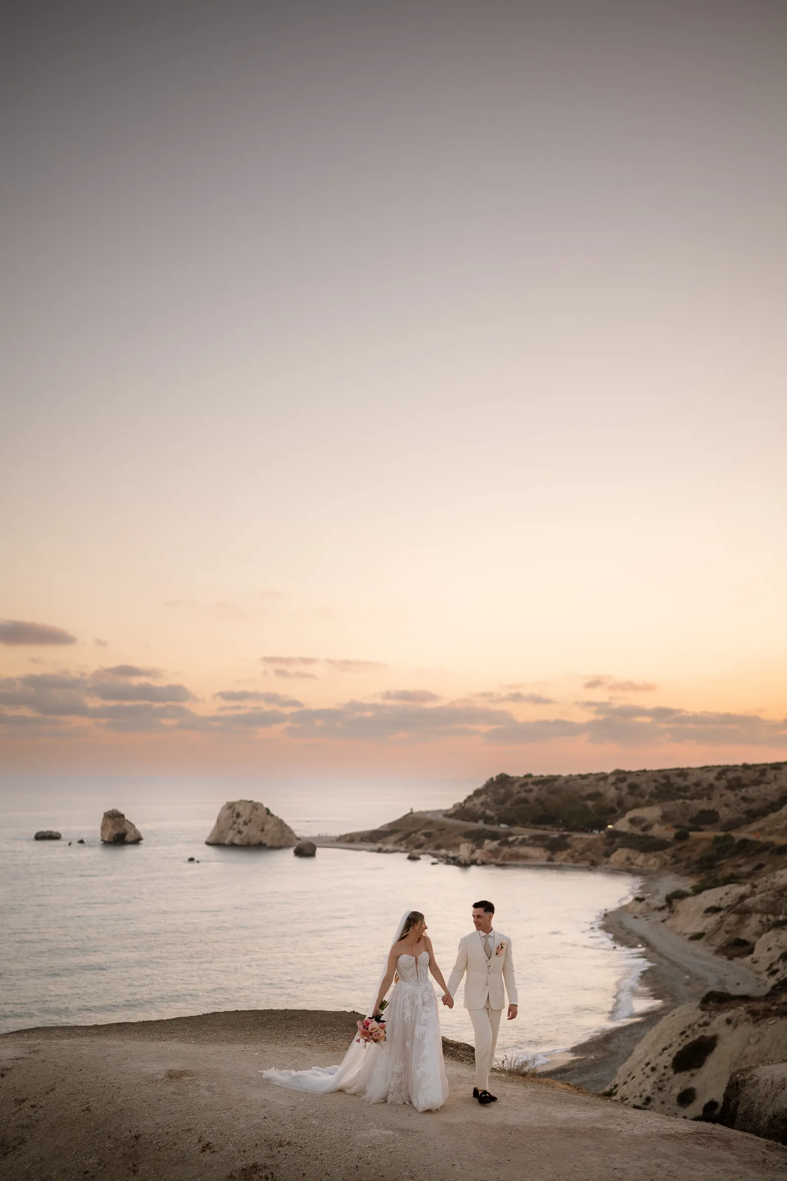 Frank and Franziska beneath a wide pastel sky on the headland at Aphrodite's Rock, the sea rock and bay stretching out below, near Paphos, Cyprus.