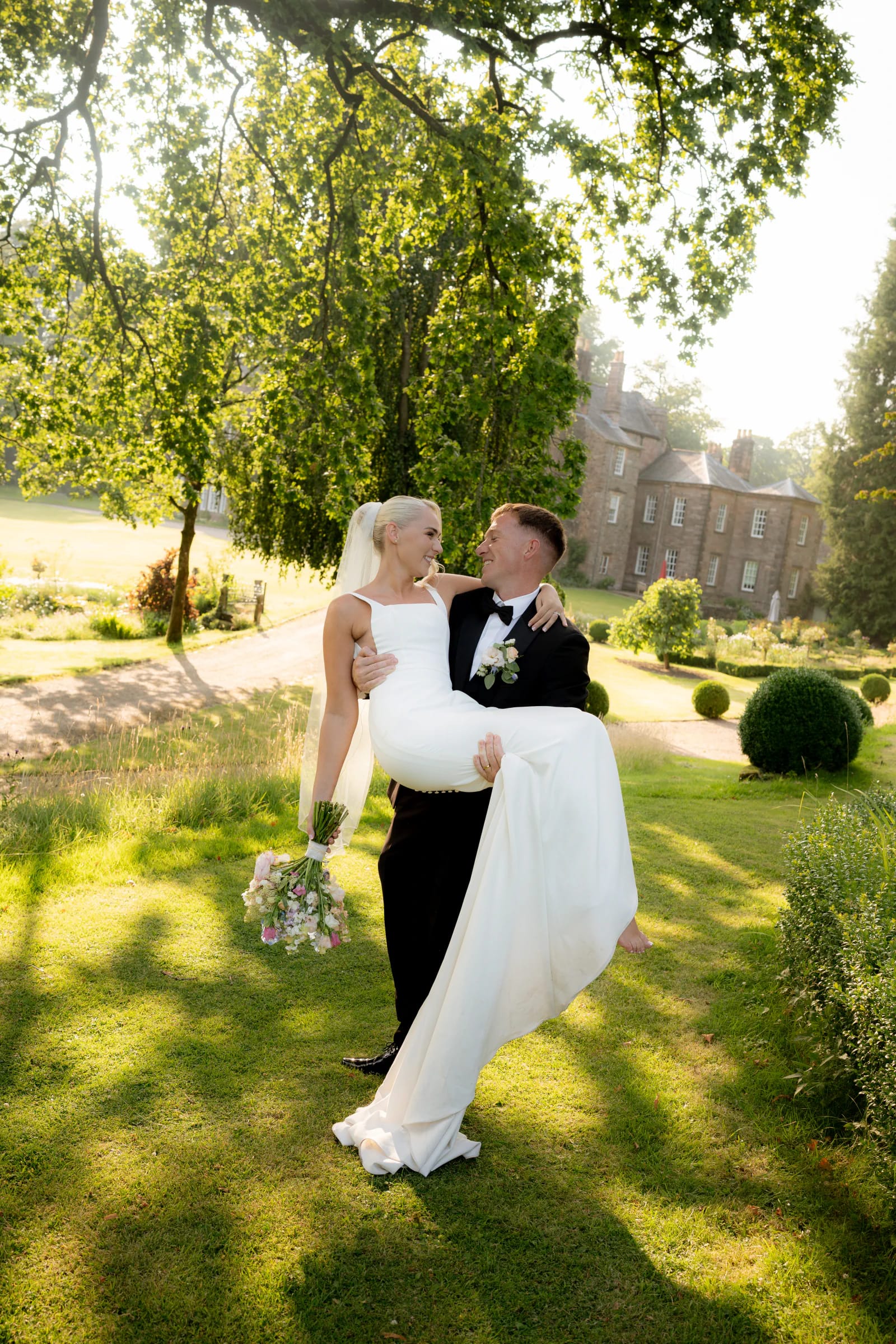 Josh carrying Ellie through the sunlit gardens with Browsholme Hall behind them