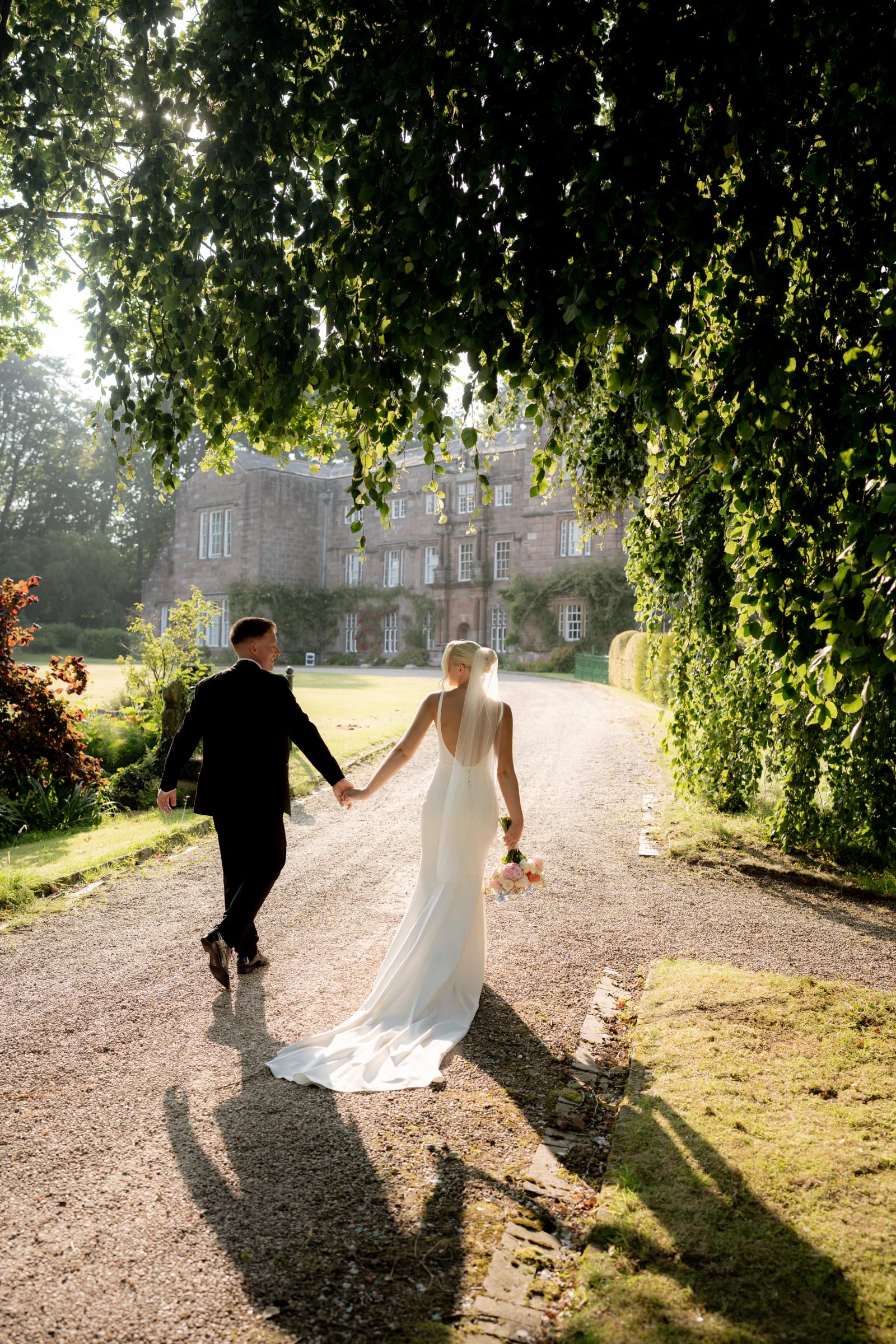 Josh and Ellie walking hand in hand towards Browsholme Hall beneath the trees in the evening light