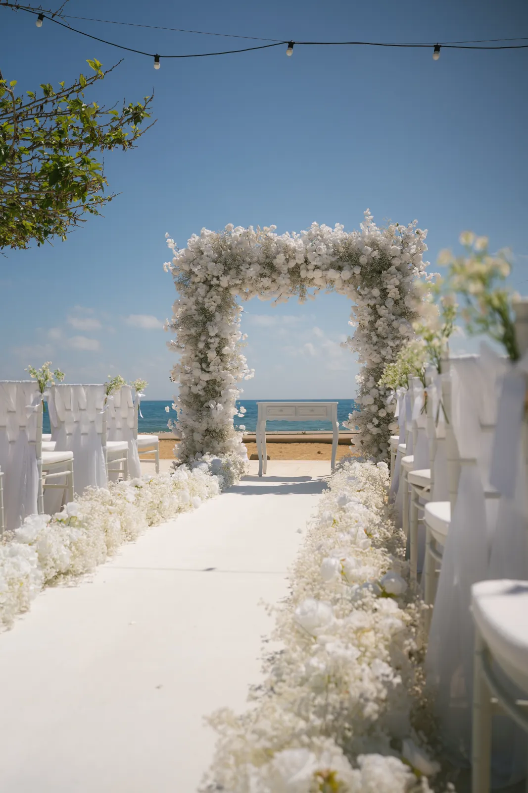 The ceremony aisle and white floral arch framed by festoon lights and the sea at Coral Residences, Paphos.