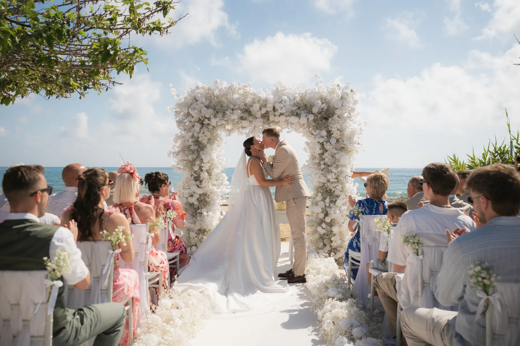 First kiss beneath a white floral arch with the sea behind at Coral Residences, Paphos, Cyprus