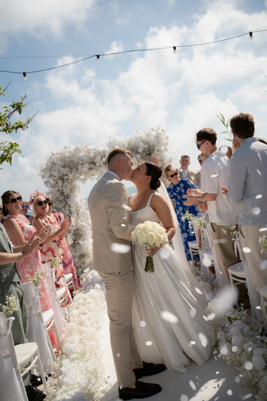 Sian and Aaron sharing their first kiss under the white floral arch as confetti falls at Coral Residences, Paphos.