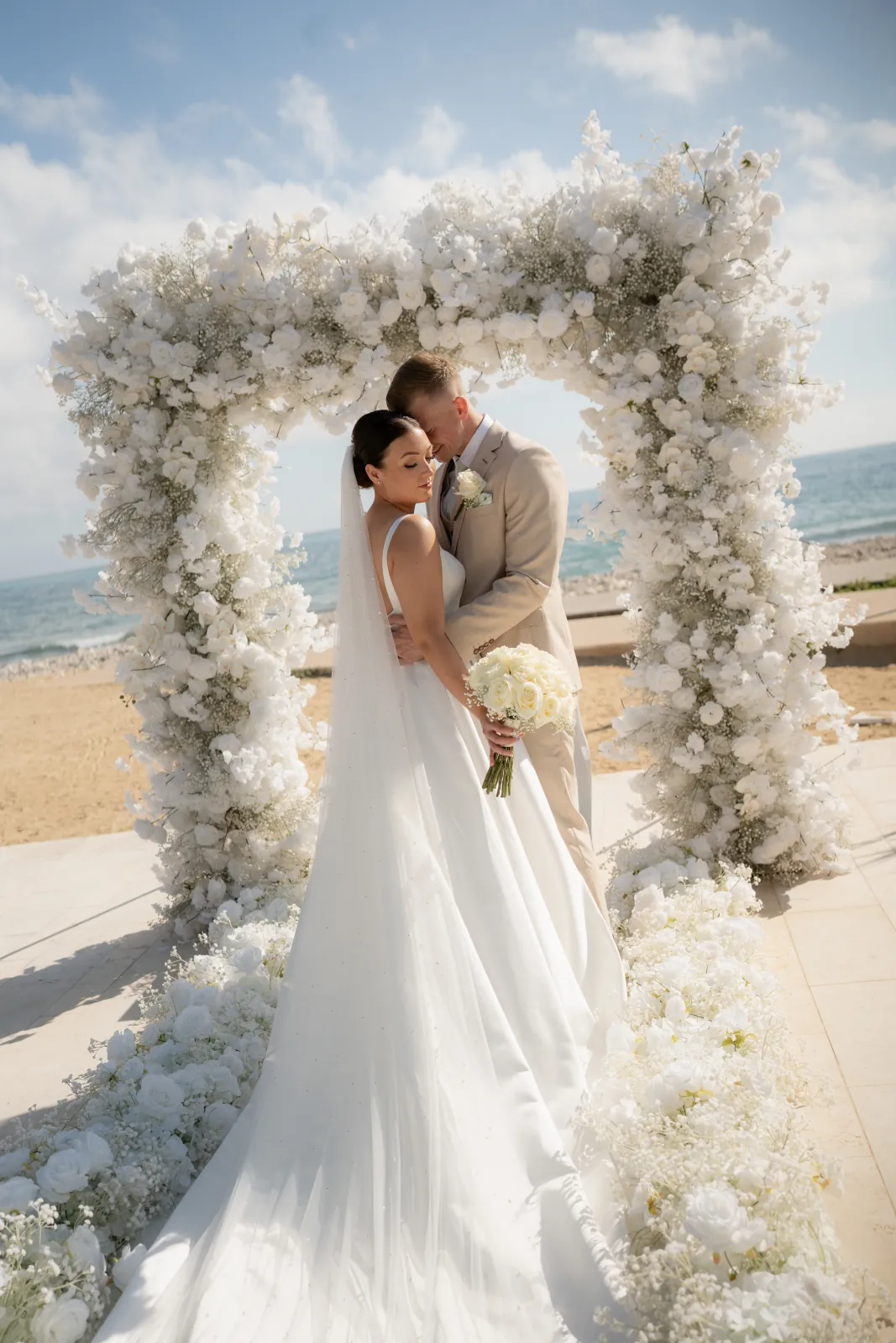 Aaron kissing Sian's temple beneath the white floral arch with the sea behind at Coral Residences, Paphos.