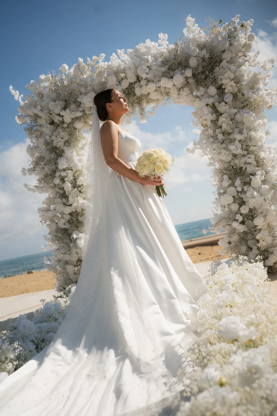 Sian alone beneath the white floral arch holding her ivory bouquet at Coral Residences, Paphos, Cyprus.